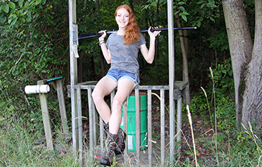 Girl with red hair sits on a fence with a skeet shooting rifle over her shoulders