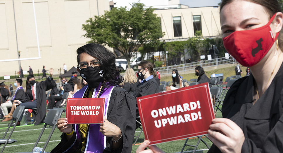 Two students in robes and masks sit at the 2021 commencement ceremony. The student on the left holds a sign that says "tomorrow the world." The student on the right holds a sign that says "Onward! Upward!"